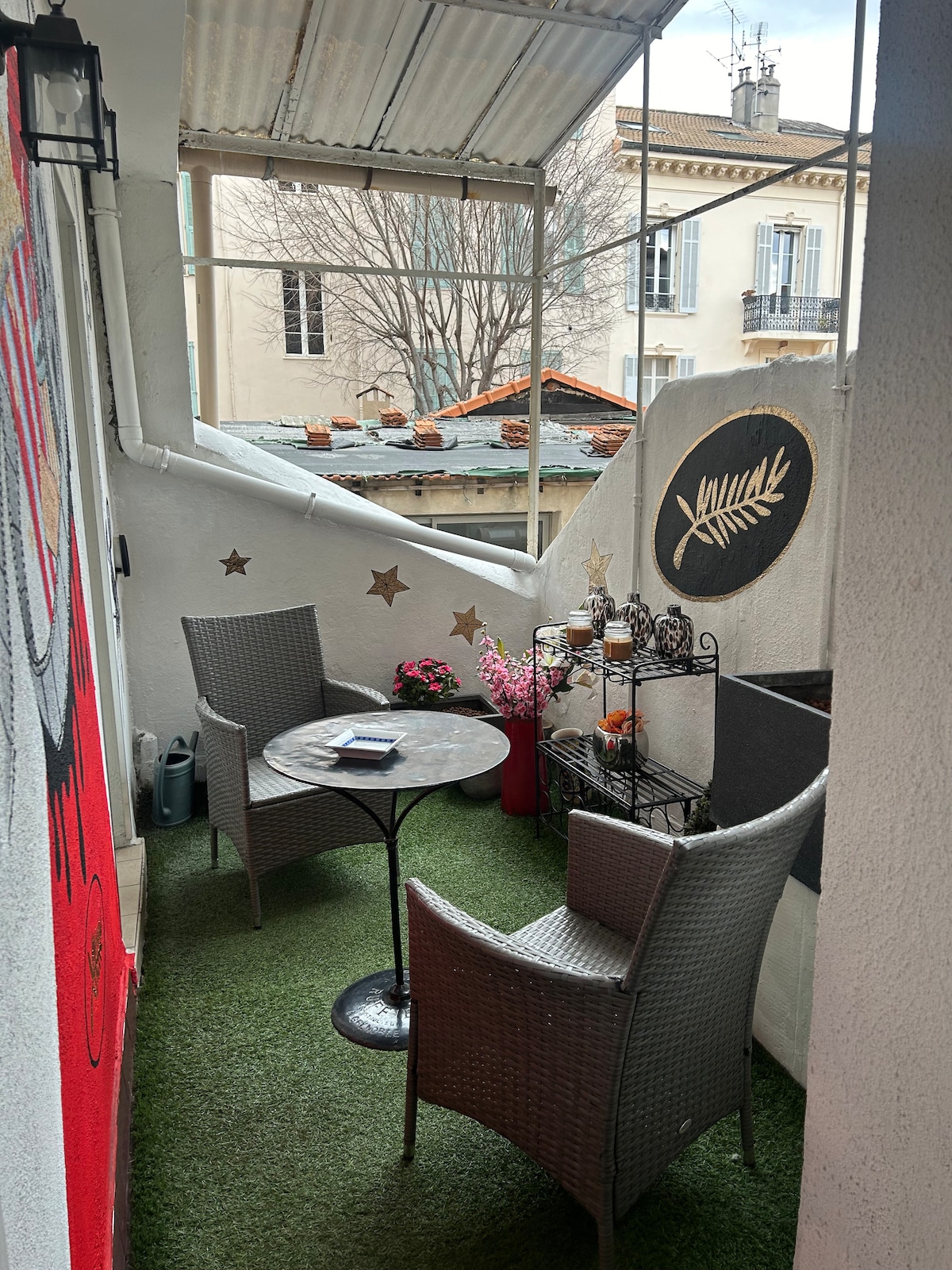 A charming balcony is shown, featuring two wicker chairs around a small table. Decorative elements include potted plants and a wall-mounted shelf with ornamental features. The flooring is carpeted with synthetic grass, creating a relaxing outdoor space with walls adorned with subtle designs.