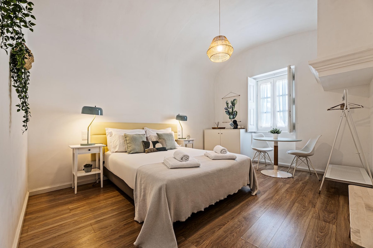 A cozy bedroom features a spacious bed with light linens, complemented by decorative pillows. A minimalist design incorporates wooden flooring and indoor plants. A small table with two chairs is present, alongside a bedside table and lamps, while natural light enters through the window.