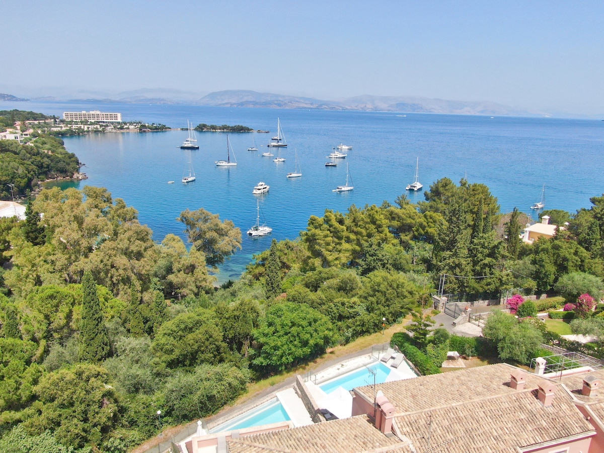 A panoramic view highlights the tranquil blue waters of Kommeno bay, dotted with anchored sailboats. Lush greenery frames the scene, while the coastline is visible in the distance, showcasing the serene beauty of the surrounding landscape and the inviting infinity pool below.