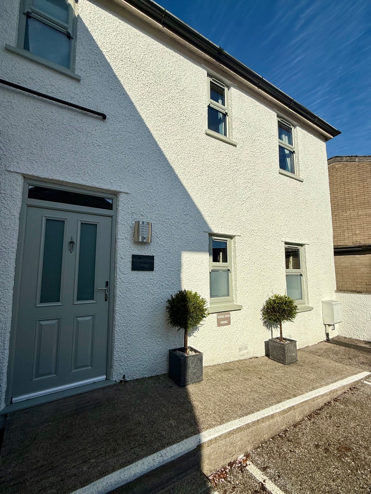 The exterior of the cottage is depicted, showcasing a clean, white facade complemented by several windows. The entrance features a light green door, framed by two potted plants. A paved pathway leads up to the door, and a parking area is visible to the side.