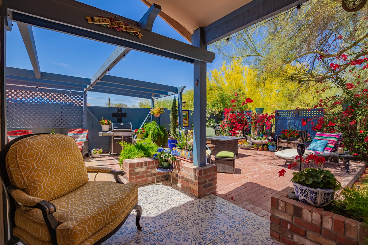 An outdoor patio space is highlighted by a patterned tile floor, surrounded by vibrant flowering plants. Comfortable seating is present, with sunlight filtering through a trellis. A gas grill is positioned for outdoor cooking, creating an inviting space for relaxation and entertainment.