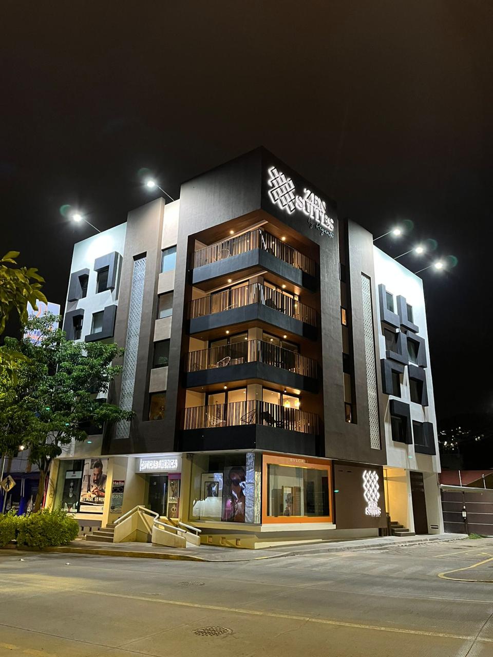 The modern building features a sleek facade with multiple balconies and illuminated signage. Its design showcases a blend of dark and light materials, with large windows allowing visibility into the interior spaces. Surrounding greenery adds a touch of nature to the urban setting.