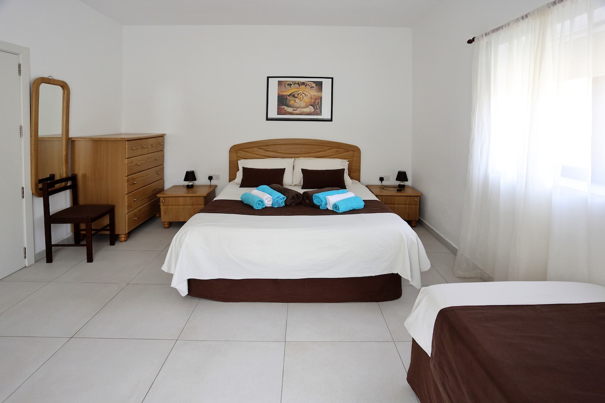 The spacious bedroom features a king-size bed and a proper single bed, both adorned with light-colored linens. Two bedside tables with lamps are positioned on either side, while a wooden dresser and a chair add functionality. Natural light filters through sheer curtains.