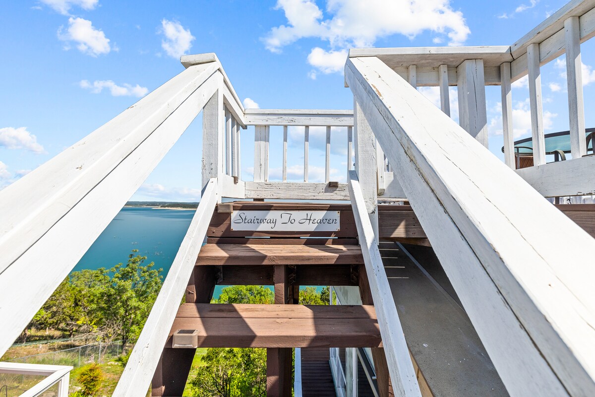 A wooden staircase leads upwards, adorned with a sign that reads 'Stairway To Heaven.' The steps are flanked by a white railing, providing a view of the expansive Canyon Lake and lush greenery in the distance under a bright blue sky.