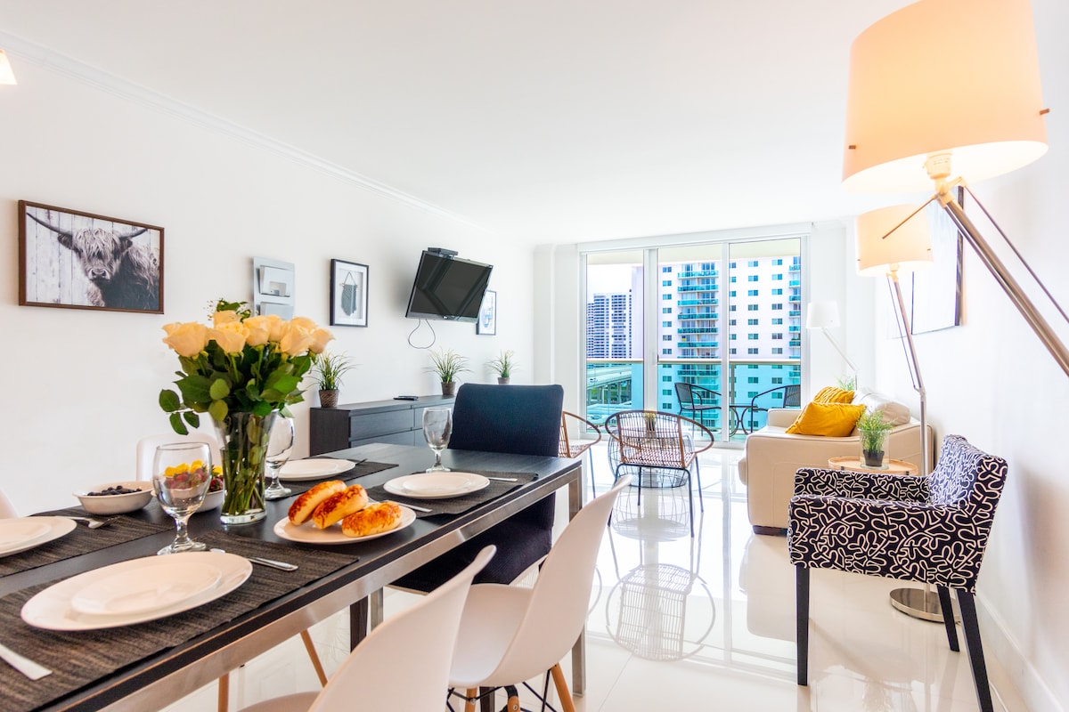 A dining area showcases a long table set for breakfast, complete with plates and glassware. Fresh roses in a vase are positioned at the center. In the background, a comfortable seating area is visible with large windows allowing natural light and city views.