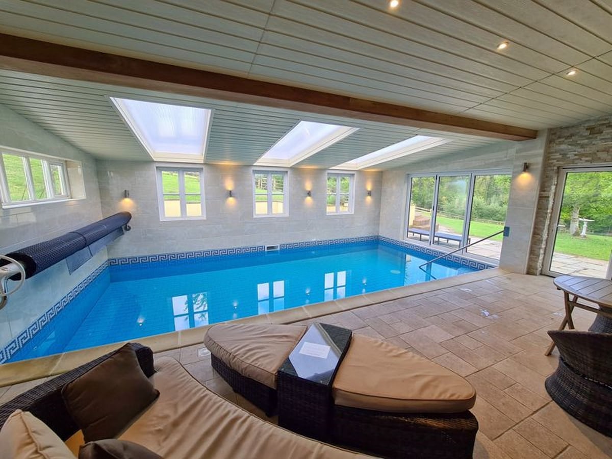 An indoor heated swimming pool is surrounded by large windows, allowing natural light to fill the space. Comfortable lounge chairs are positioned beside the pool, providing a relaxing area. The pool area opens directly to a grassy outdoor space, inviting guests to enjoy both indoor and outdoor leisure.