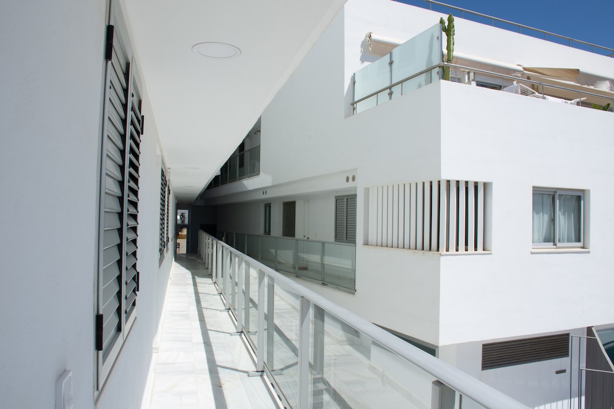 A bright corridor features a smooth tile floor and a glass railing along one side. Large windows are positioned intermittently along the white exterior walls, allowing natural light to enhance the space. Louvered shutters are present on several windows, adding to the design.