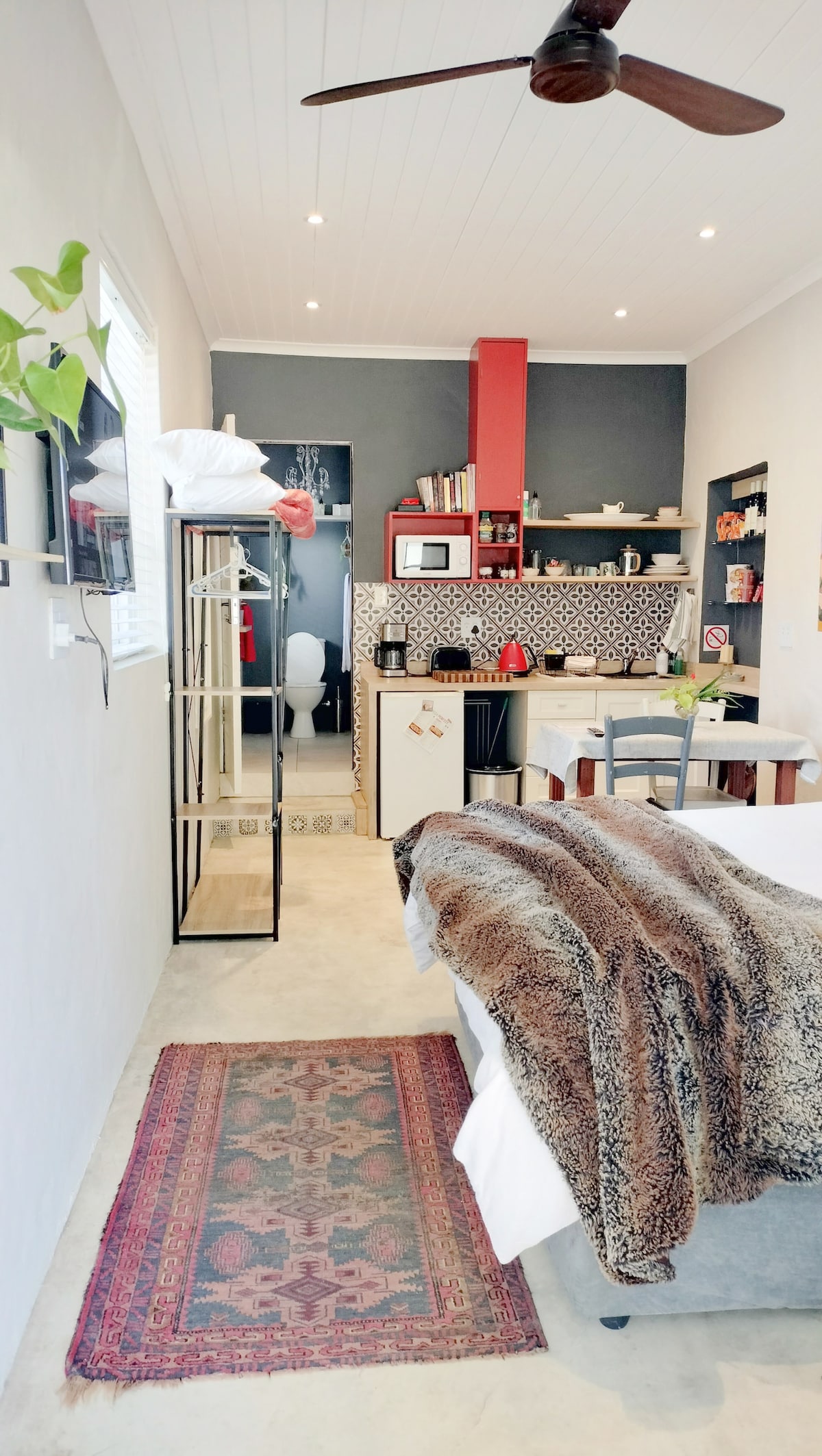 The cozy interior features a spacious bed adorned with a textured blanket and a vintage rug underfoot. A kitchenette is visible along the wall, equipped with a microwave and ample storage. Natural light enters through a window, enhancing the modern design.