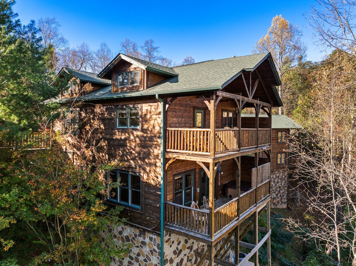 The exterior view of the two-level cabin showcases a wooden facade and multiple balconies adorned with railings. Surrounding trees blend with the natural landscape, and a stone foundation adds texture to the structure. The clear sky provides a backdrop for this serene mountain retreat.