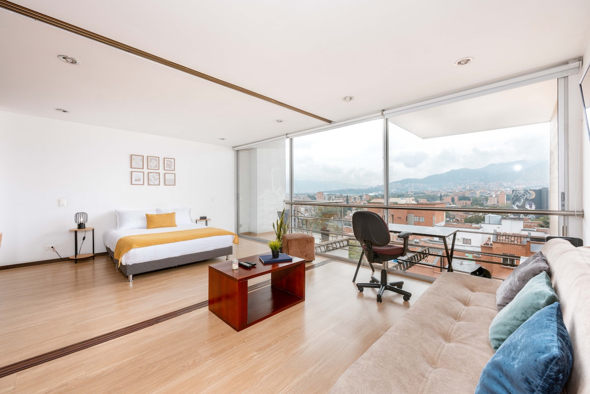 A spacious studio features large windows providing ample natural light and a panoramic view of the city. A comfortable sofa, a simple coffee table, and a work desk are arranged in the open area, while a neatly made bed offers a cozy sleeping corner.