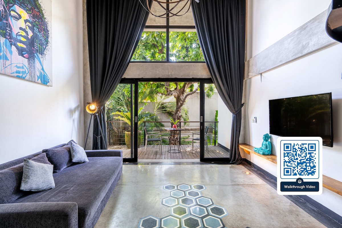 A spacious living area features a comfortable gray sofa positioned under large windows, allowing natural light to enter. Dark curtains frame the windows, and an accent wall with artwork adds character. A television is mounted on the wall opposite, while a terrace offers views of greenery.