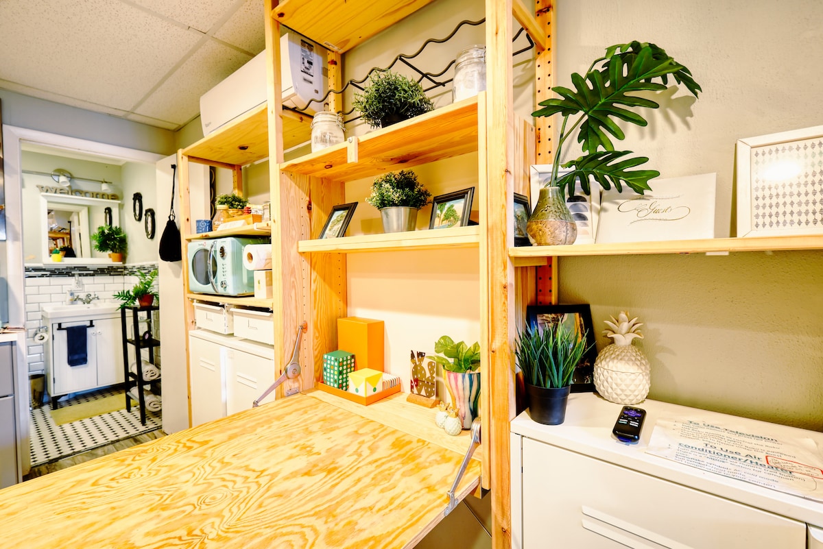A wooden folding table is seen with wooden shelving above it, displaying decorative plants and small framed pictures. To the left, a kitchenette area is visible with white cabinetry and a retro microwave. The decor features hints of greenery, adding a welcoming touch.