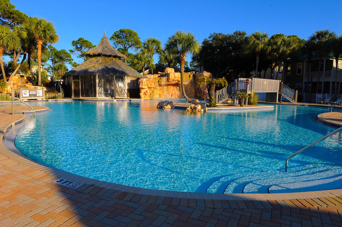 A spacious tropical pool is surrounded by lush palm trees and a charming thatched-roof structure. The inviting water reflects the clear blue sky, while rock formations and gentle waterfalls enhance the natural beauty of the area. Lounge chairs are visible along the pool's edge.