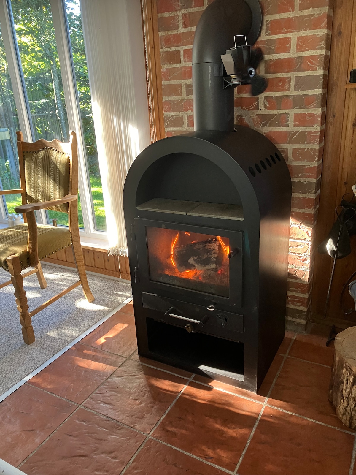 A black wood stove is positioned against a brick wall, providing warmth to the room. Sunlight filters through large windows, casting light onto the sleek surface of the stove. A wooden chair is seen nearby, enhancing the cozy ambiance of the space.