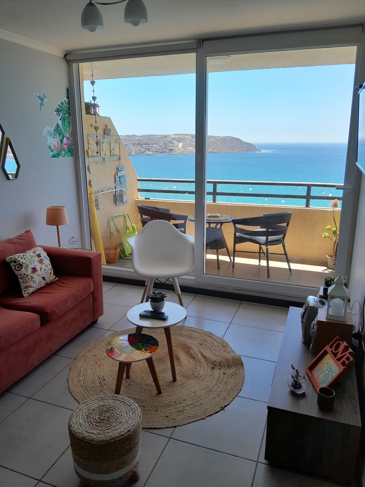 A cozy living space is highlighted by a red sofa and a small, round coffee table. Floor-to-ceiling glass doors provide a panoramic view of the ocean and the surrounding landscape. A decorative rug adds texture to the tiled floor, enhancing the inviting ambiance.