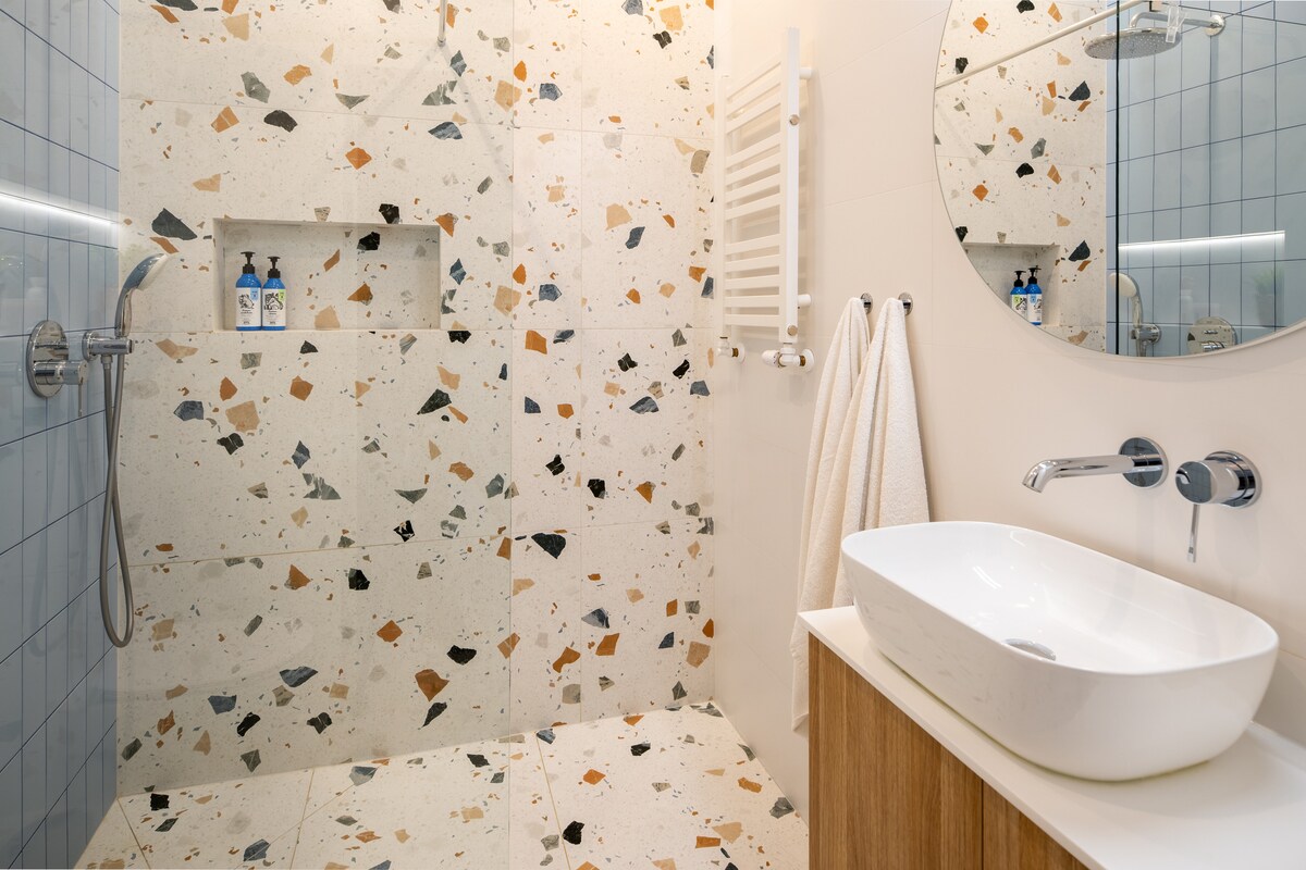 A modern bathroom features a spacious walk-in shower with a textured stone wall, showcasing unique color flecks. A circular mirror above a minimalist sink complements the wooden cabinet below. Towels are neatly hung, and personal care products are placed on the shower shelf.