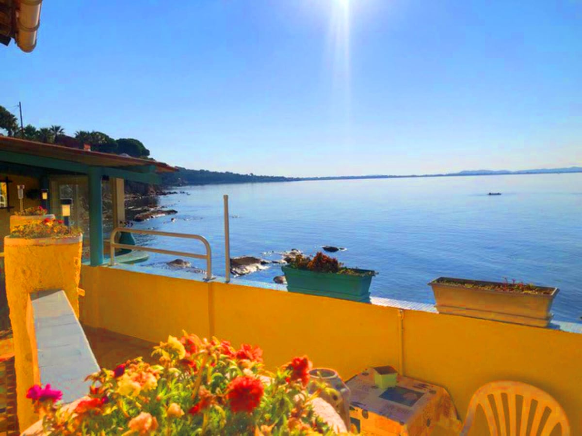 A serene view of the calm sea is captured, with boats visible in the distance. Bright flowers adorn the terrace, complementing the inviting atmosphere. The sun shines brightly, reflecting off the water, while the coastline stretches along the horizon.