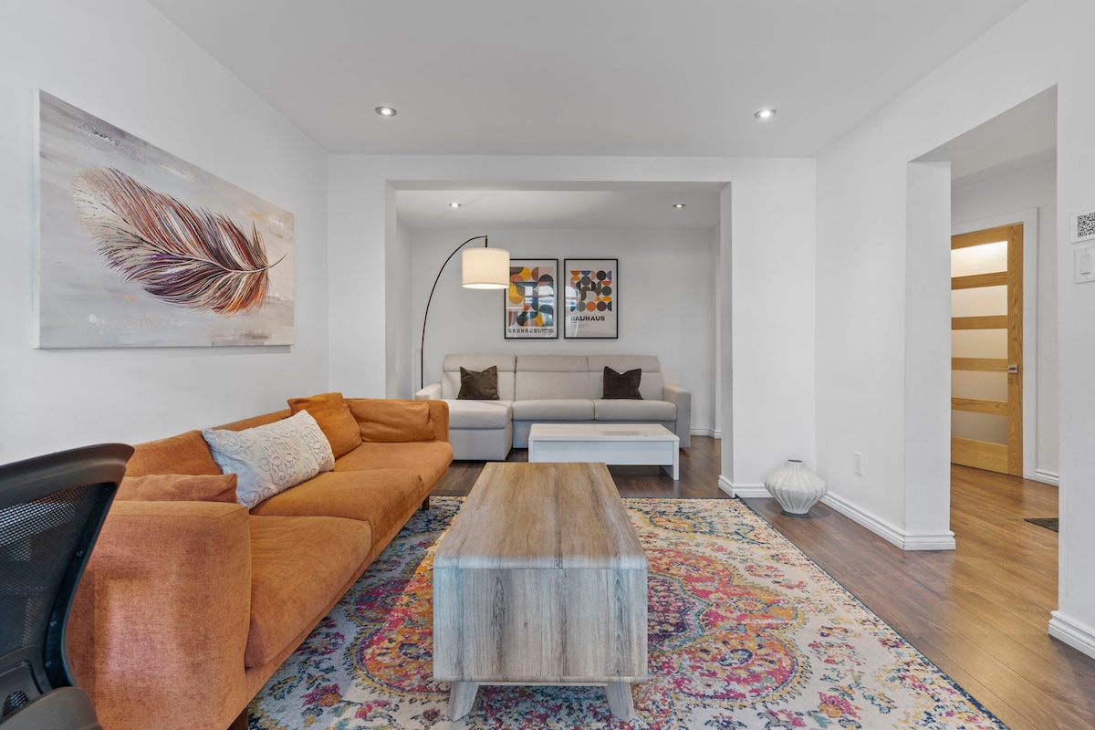 A contemporary living area features a plush orange sofa and a light-colored sectional couch, complemented by a wooden coffee table. Colorful artwork adorns the walls, and a patterned rug adds warmth to the space. Soft lighting is provided by a floor lamp and recessed fixtures.