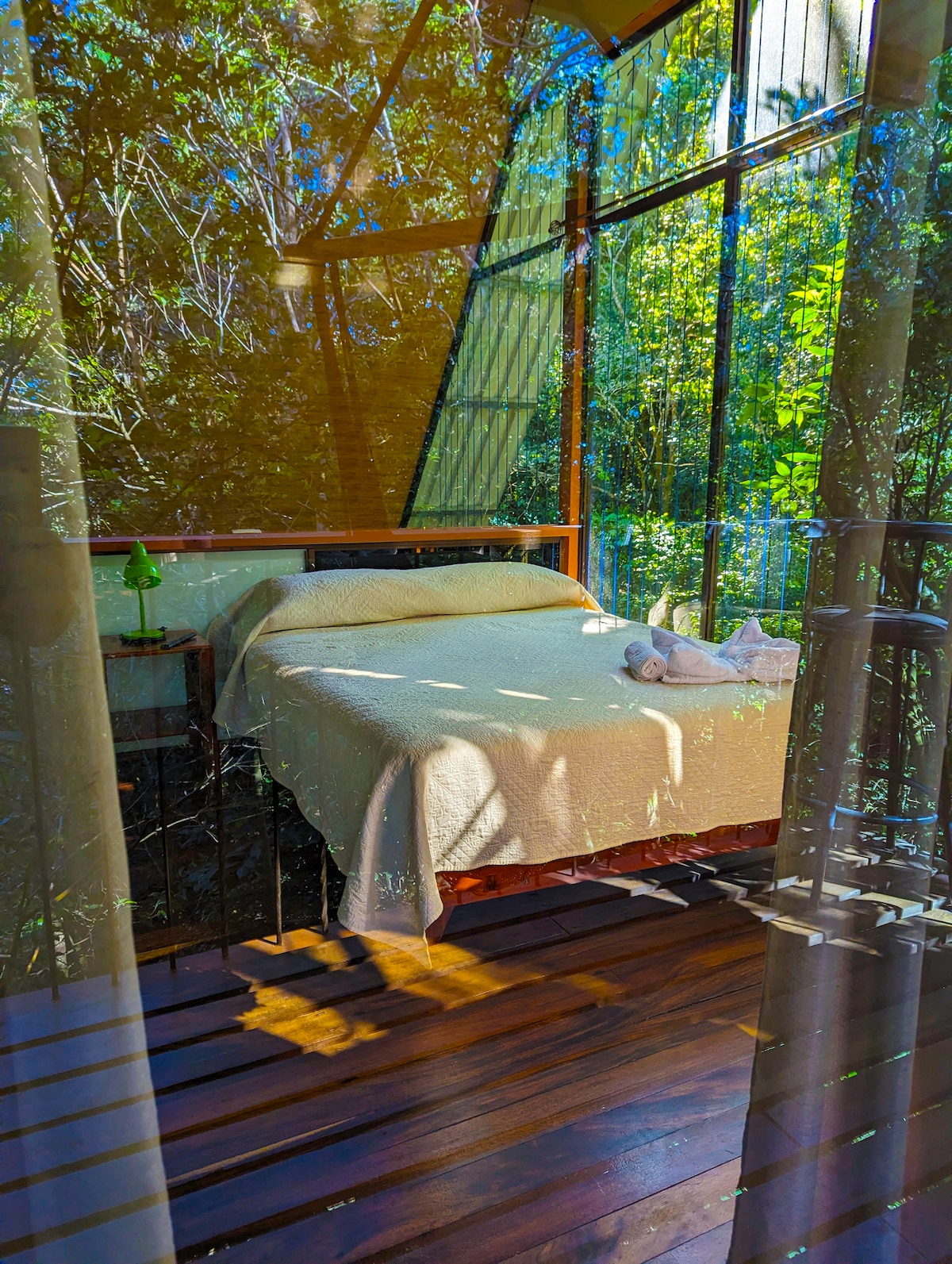 A well-lit sleeping area features a spacious bed draped with a soft blanket, set against a backdrop of greenery seen through large windows. Natural light filters in, creating a serene atmosphere. A bedside table holds a small lamp, enhancing the cozy feel.