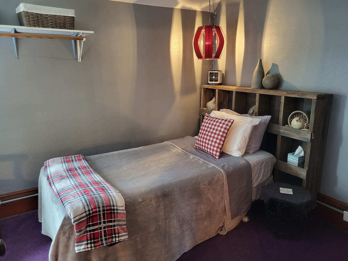 A single bed is positioned against a textured wall, featuring a plaid throw. A wooden headboard displays decorative items, and a small shelf is mounted above. A red pendant light casts soft shadows, contributing to the cozy ambiance of the space.