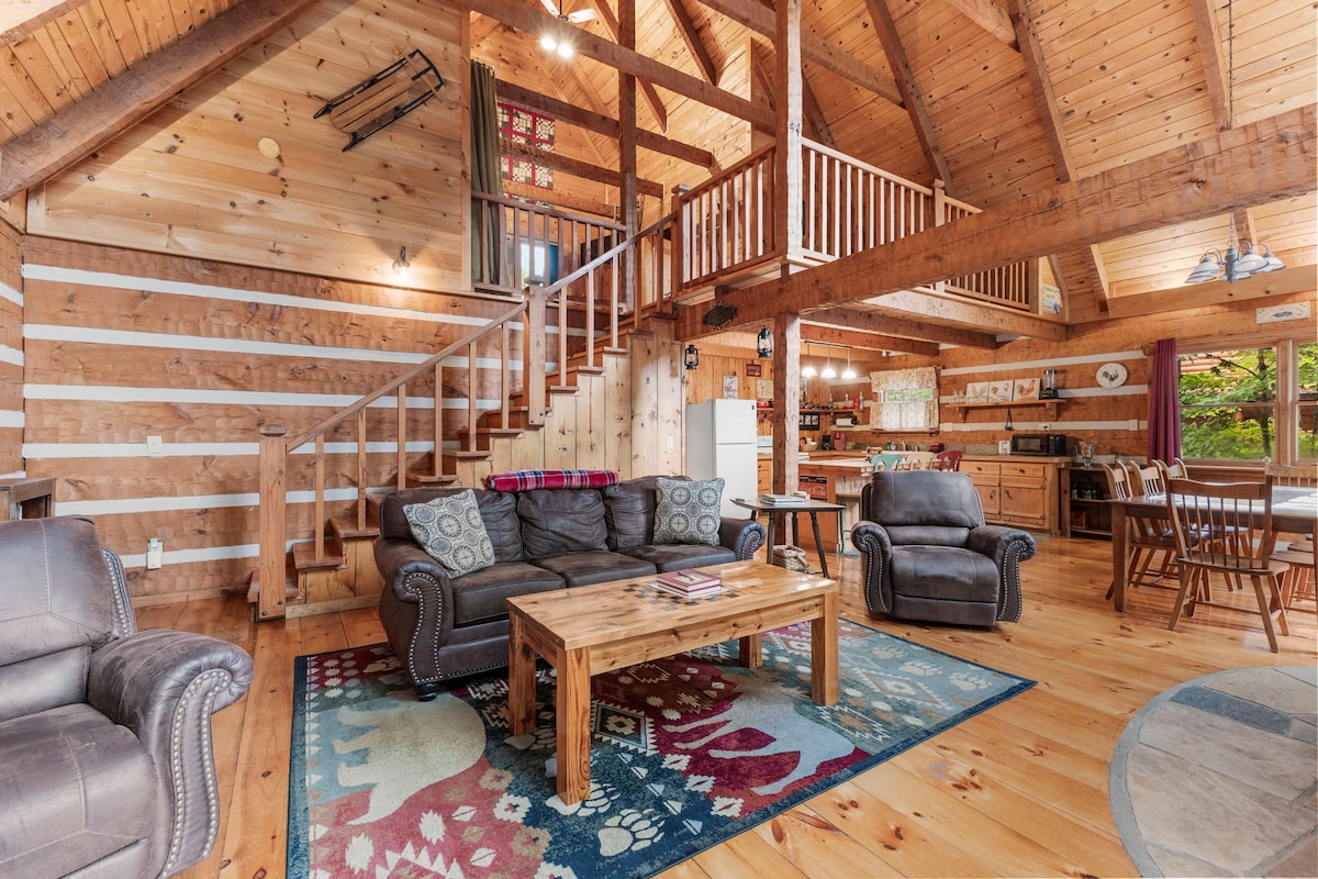 A spacious living area features high wooden ceilings and exposed beams, enhancing the cabin ambiance. Comfortable seating includes a brown couch and two recliners arranged around a wooden coffee table on a decorative rug. A staircase leads to the loft, while a fully equipped kitchen is visible in the background.