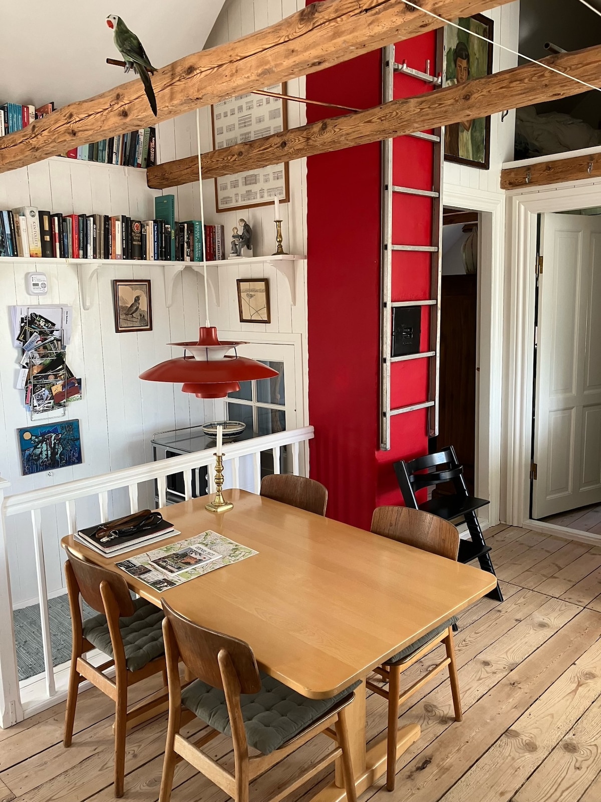 A dining area is displayed with a wooden table surrounded by four chairs. A retro lamp hangs above the table, while lined bookshelves and framed artworks adorn the walls. A red column and an ornamental ladder add visual interest, complemented by a bird perched nearby.