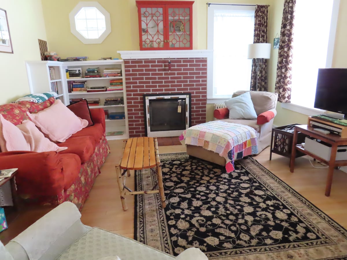 A cozy living room features a red couch adorned with soft pillows and a crocheted throw over a beige armchair. A wooden coffee table sits on a patterned rug, while bookshelves and a television add to the inviting space. Natural light filters through the windows, enhancing the warmth.