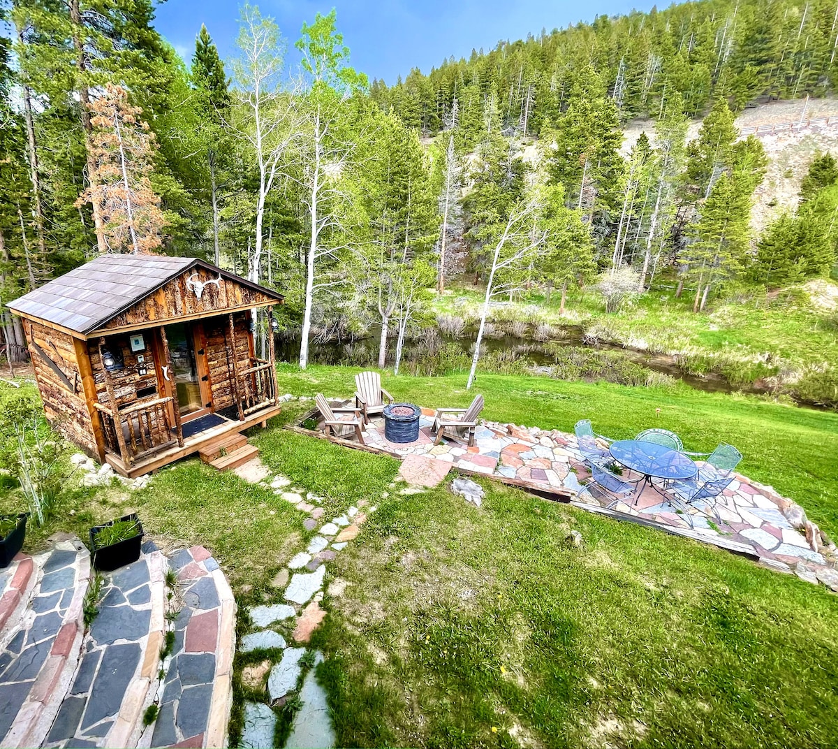 A rustic log cabin sauna is situated in the backyard, surrounded by lush greenery and trees. A stone patio features a propane fire pit and various seating options, including colorful chairs and lounge spots, all overlooking a tranquil creek lined by a grassy area.