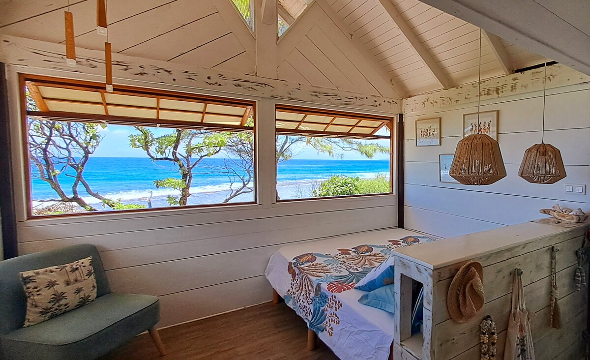 A bright and airy room features large windows offering views of the ocean and surrounding trees. A cozy bed adorned with colorful linens is complemented by natural wood accents and woven light fixtures, creating a serene and inviting space.