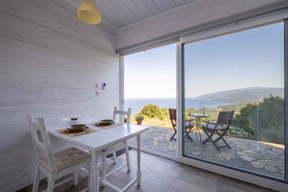 A bright dining area with a white table and chairs is positioned near large glass sliders, offering a panoramic view of the sea and mountains. A small potted plant adorns the table, and outdoor seating is visible on the stone terrace just beyond.