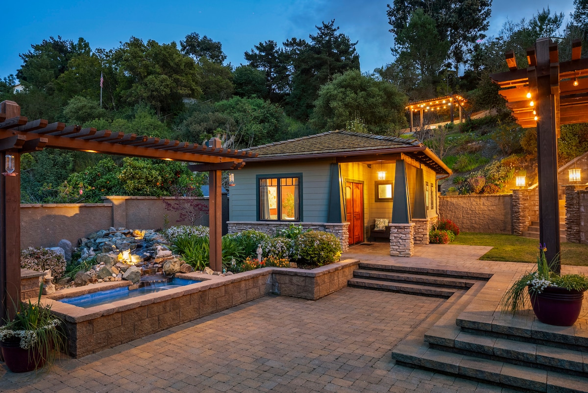 The exterior of the cozy Garden House is highlighted by warm lighting and landscaped gardens. A small water feature adds tranquility, while stone steps lead to an inviting entrance. The surrounding trees and plantings enhance the natural setting, creating a peaceful atmosphere.