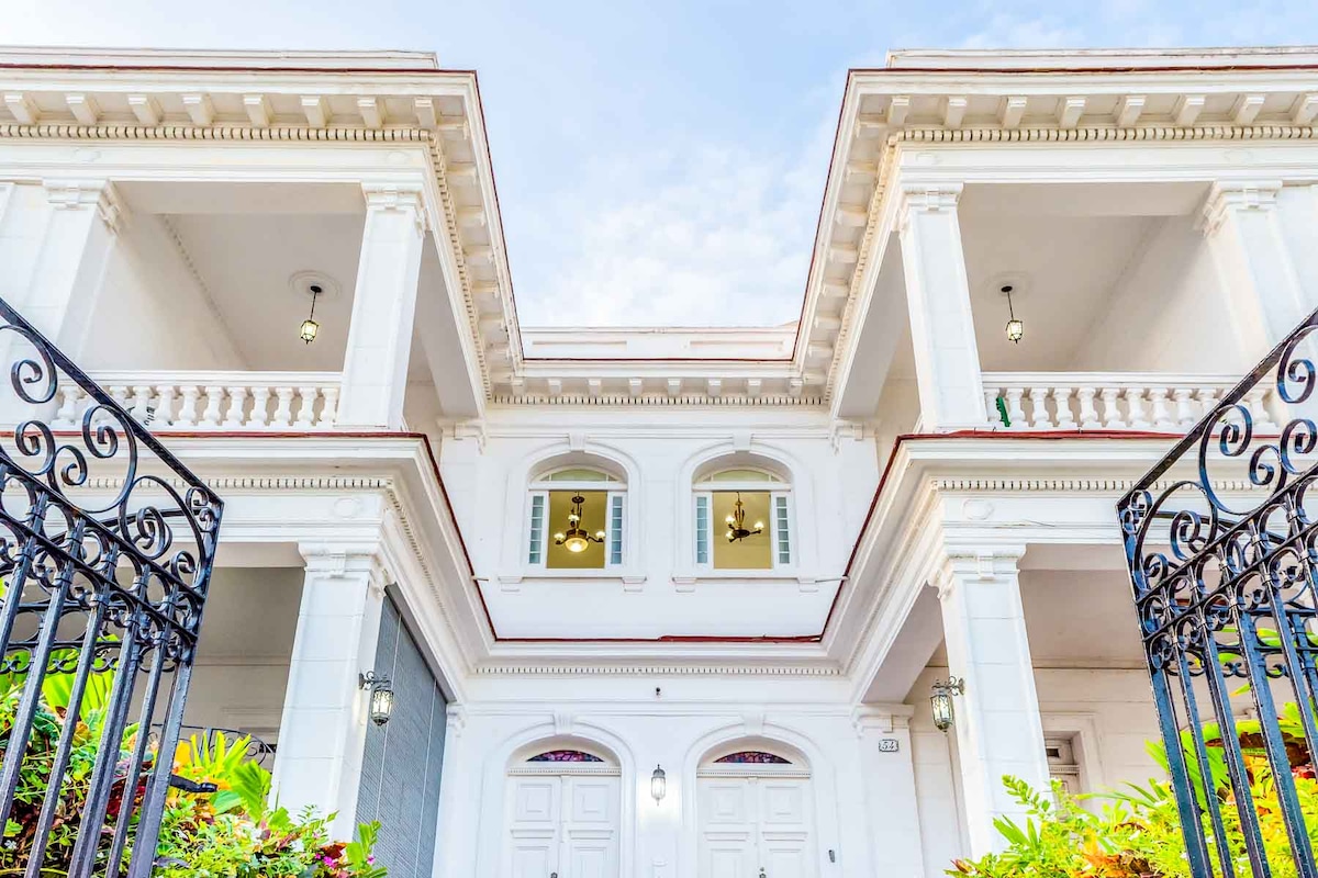 An elegant Art Deco building is showcased, featuring two symmetrical stories with classic architectural elements. The facade includes large windows and a grand entrance with double doors, framed by lush greenery and decorative wrought-iron gates.