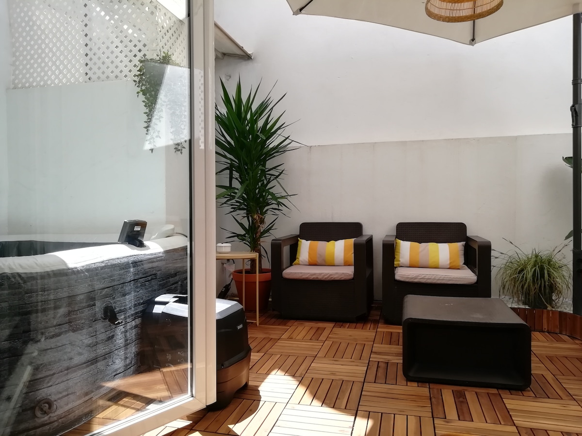 An interior terrace features two comfortable chairs with striped cushions and a low coffee table. Surrounding greenery adds a touch of nature. A hot tub is visible in the corner, and natural light filters through the glass doors.