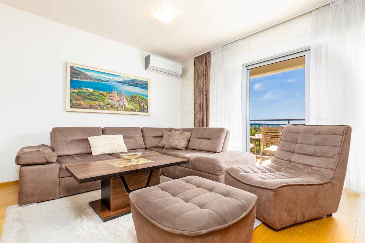 A modern living area features a sectional sofa arranged around a coffee table. Natural light floods the space through large windows, providing a view of a balcony. A vibrant artwork hangs on the wall, enhancing the room's open and inviting feel.