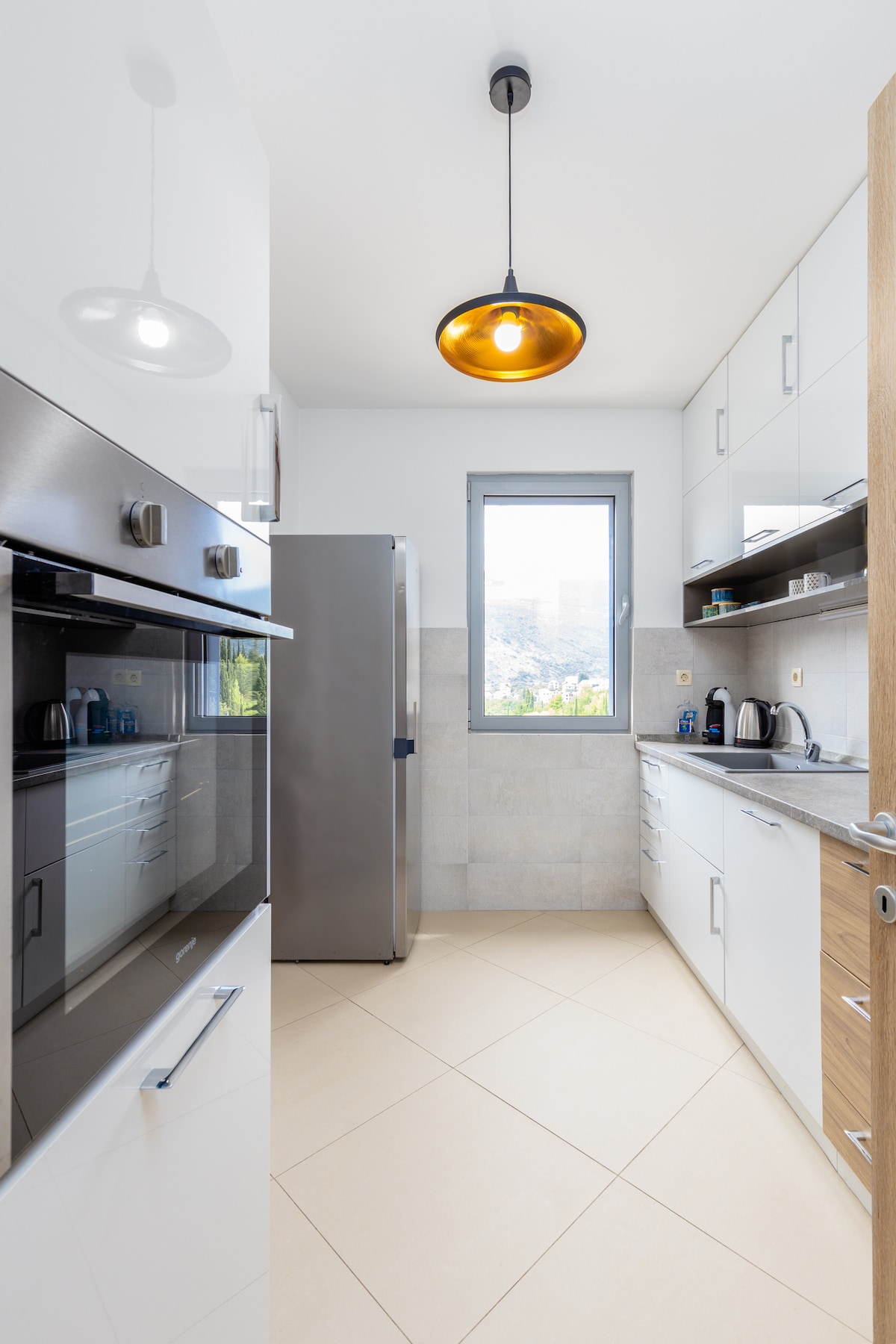 A modern kitchen displays sleek cabinetry and stainless steel appliances. A large refrigerator stands alongside built-in ovens, while a window offers natural light and views of the exterior greenery. The light fixtures above contribute to a bright and functional cooking space.