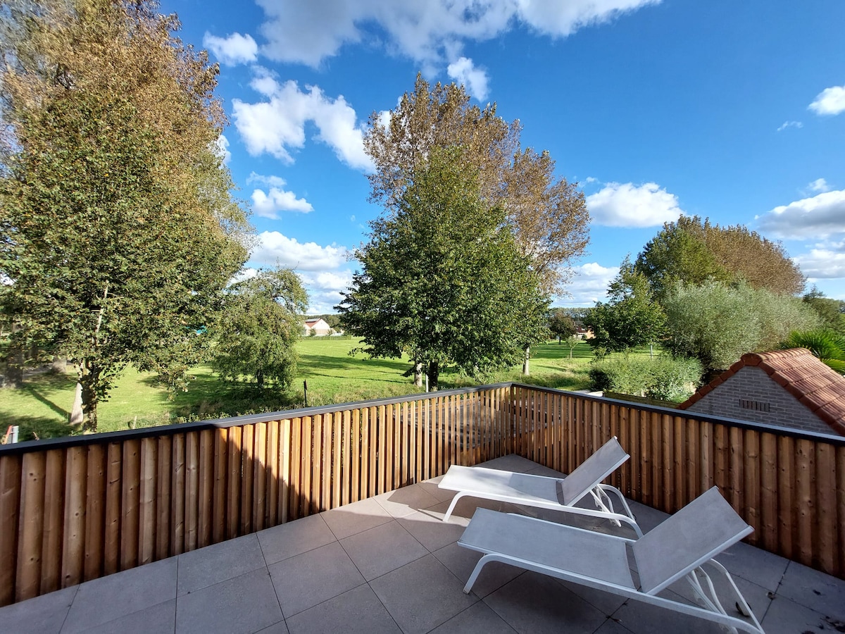 A private terrace is showcased, featuring two lounge chairs placed on a tiled surface. A wooden railing provides a view of the lush green landscape and trees under a bright blue sky with scattered clouds.