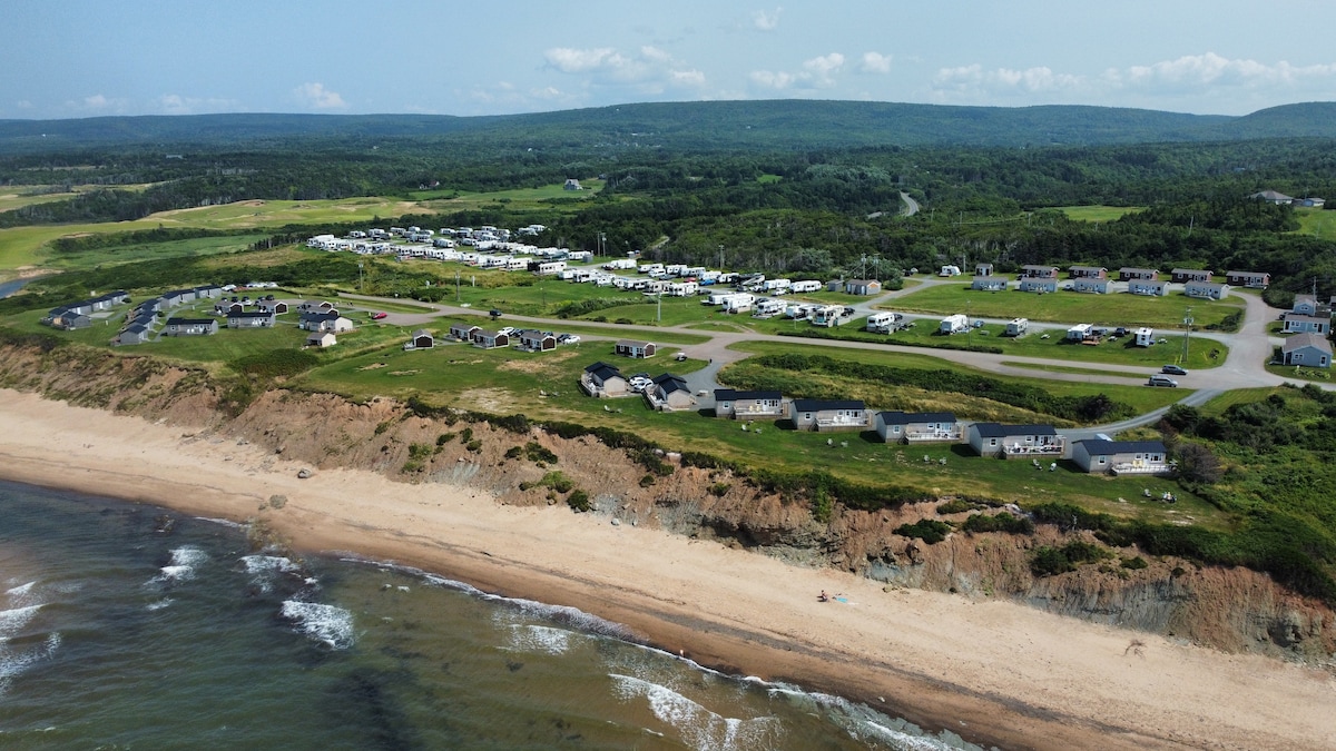 An aerial view showcases a stretch of sandy beach beside a cluster of cottages and camping sites. Expansive green fields and wooded areas provide a picturesque backdrop, while the gentle waves of the ocean lap at the shore.
