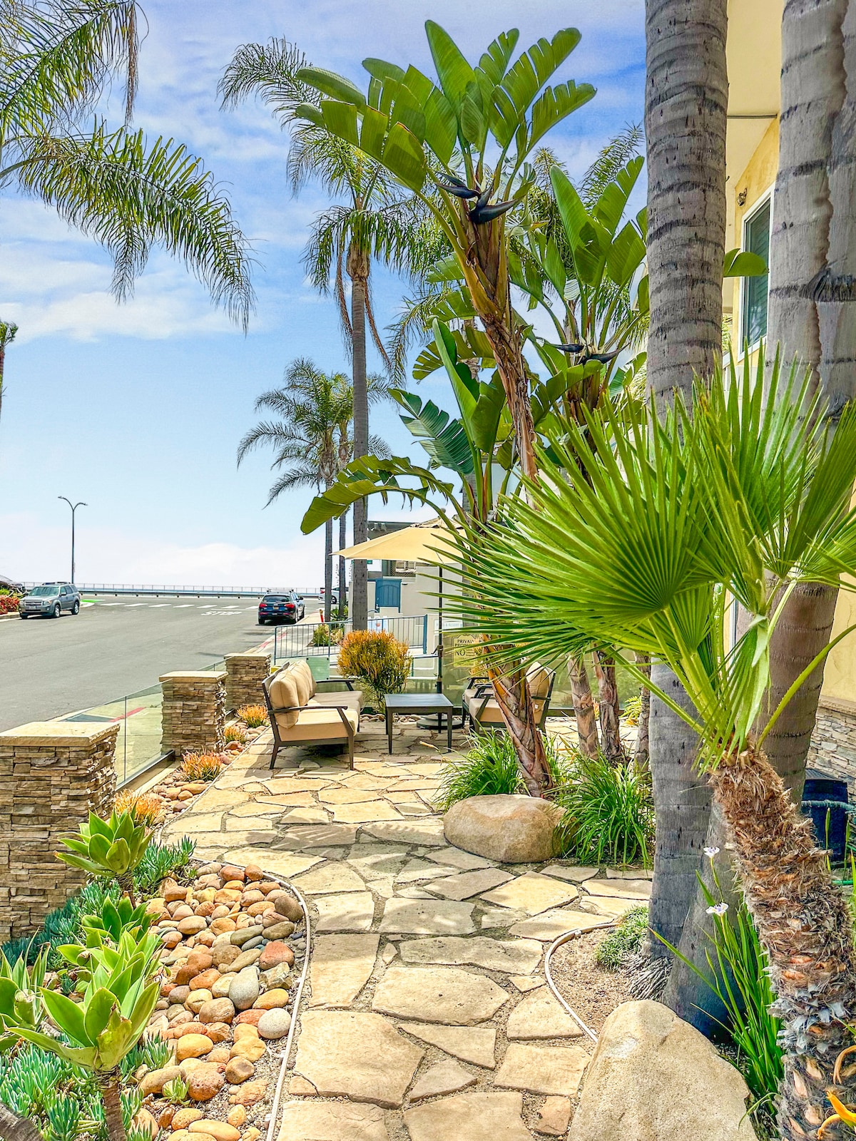 A stone pathway is lined with tropical plants and palm trees, leading to a seating area that includes two chairs and a table. The surrounding landscaping features various rocks and greenery, creating a serene environment. A glimpse of the road and distant ocean can be seen.