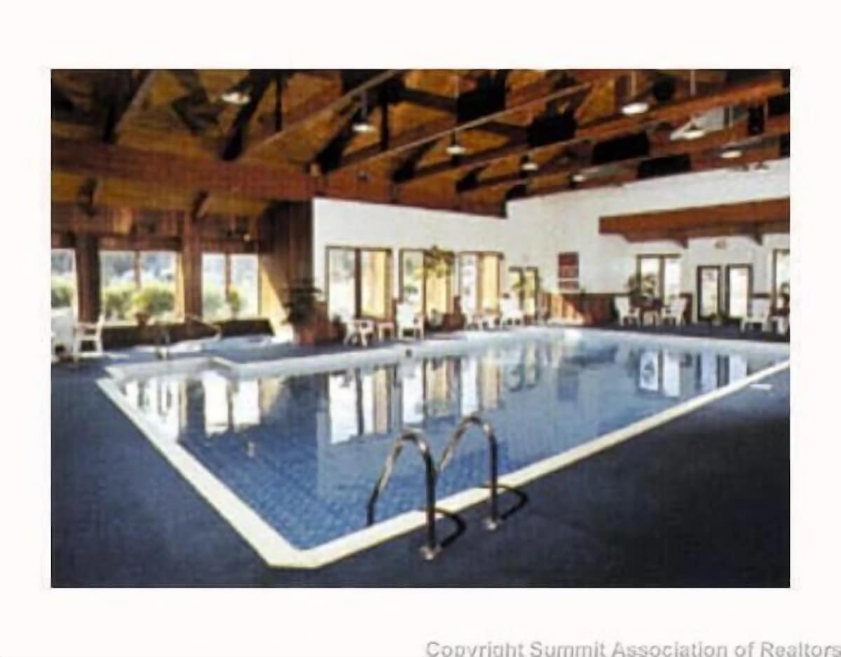 An indoor swimming pool is framed by large windows that allow natural light to fill the space. The pool area features a blue tiled surface and is surrounded by white lounge chairs. Wood beams add warmth to the high ceiling.