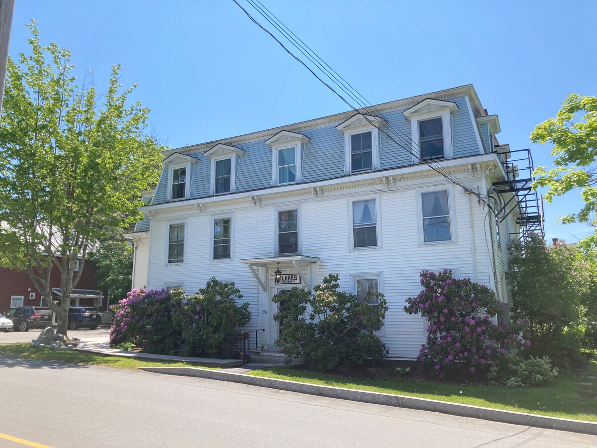 The historic building features three stories with a traditional white exterior and classic design elements. Lush green foliage and blooming flowers flank the entrance, while clear blue skies provide a bright backdrop. A welcoming atmosphere is suggested by the inviting front porch and nearby seating.