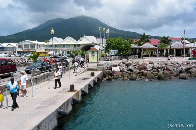 A waterfront scene features a bustling promenade alongside the water. People stroll along the path, while vehicles are parked nearby. Lush greenery and traditional architecture are visible in the background, with a prominent mountain rising under a partly cloudy sky.