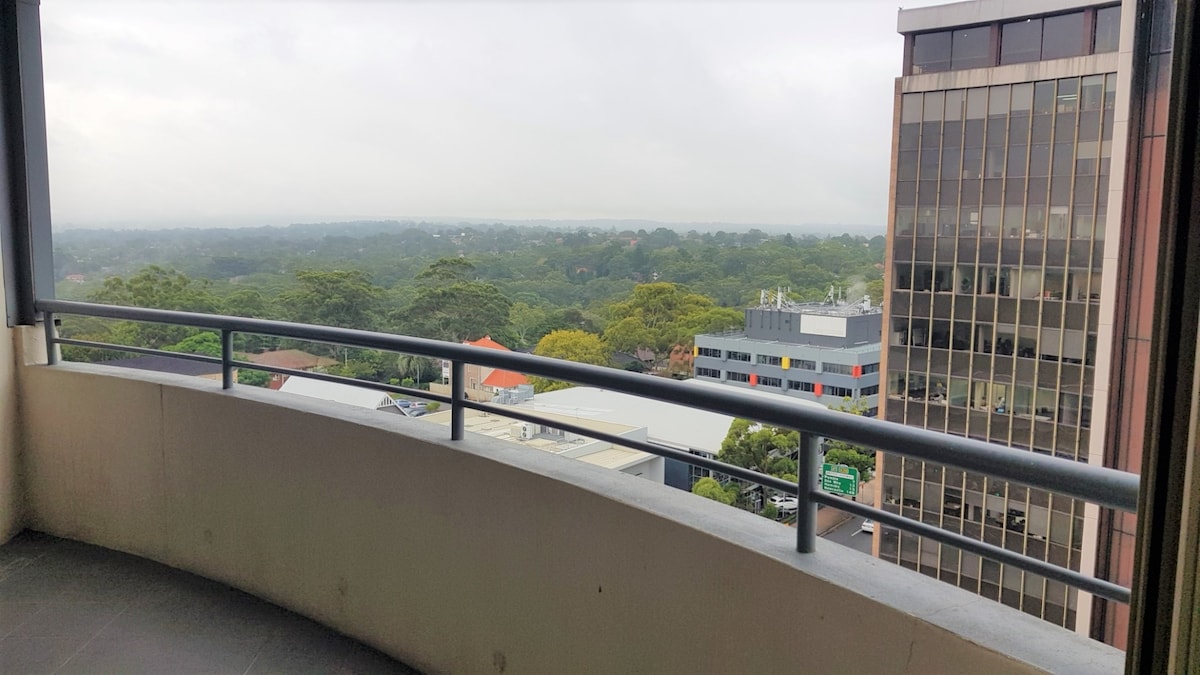 A spacious balcony showcases a view of the surrounding greenery and urban landscape. The railing provides safety while offering an unobstructed sight of the buildings and trees below, inviting moments of relaxation and enjoyment of the fresh air.