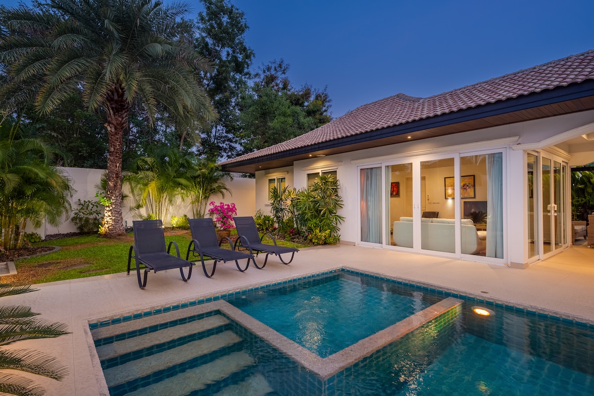 An outdoor space features a private pool surrounded by tropical greenery and a well-manicured garden. Two black sunbeds are positioned by the water, and floor-to-ceiling glass doors reflect the interior living area, creating a seamless connection between indoor and outdoor environments.