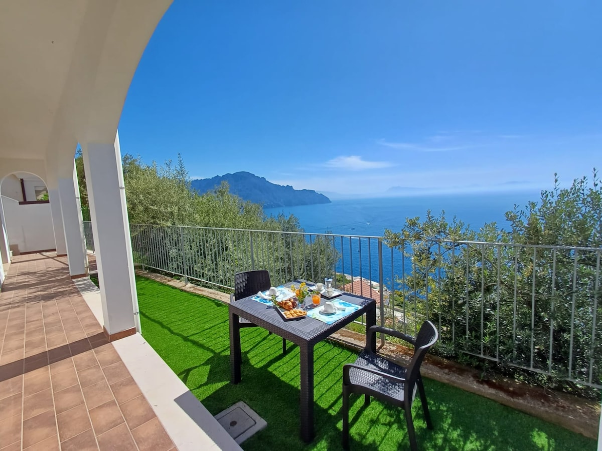 An outdoor terrace is shown, featuring a dining table set for meals with views of the coastline and the Mediterranean Sea. Lush greenery surrounds the space, and an expansive sky adds to the tranquil atmosphere. The table is neatly arranged with breakfast items, enhancing the inviting setting.
