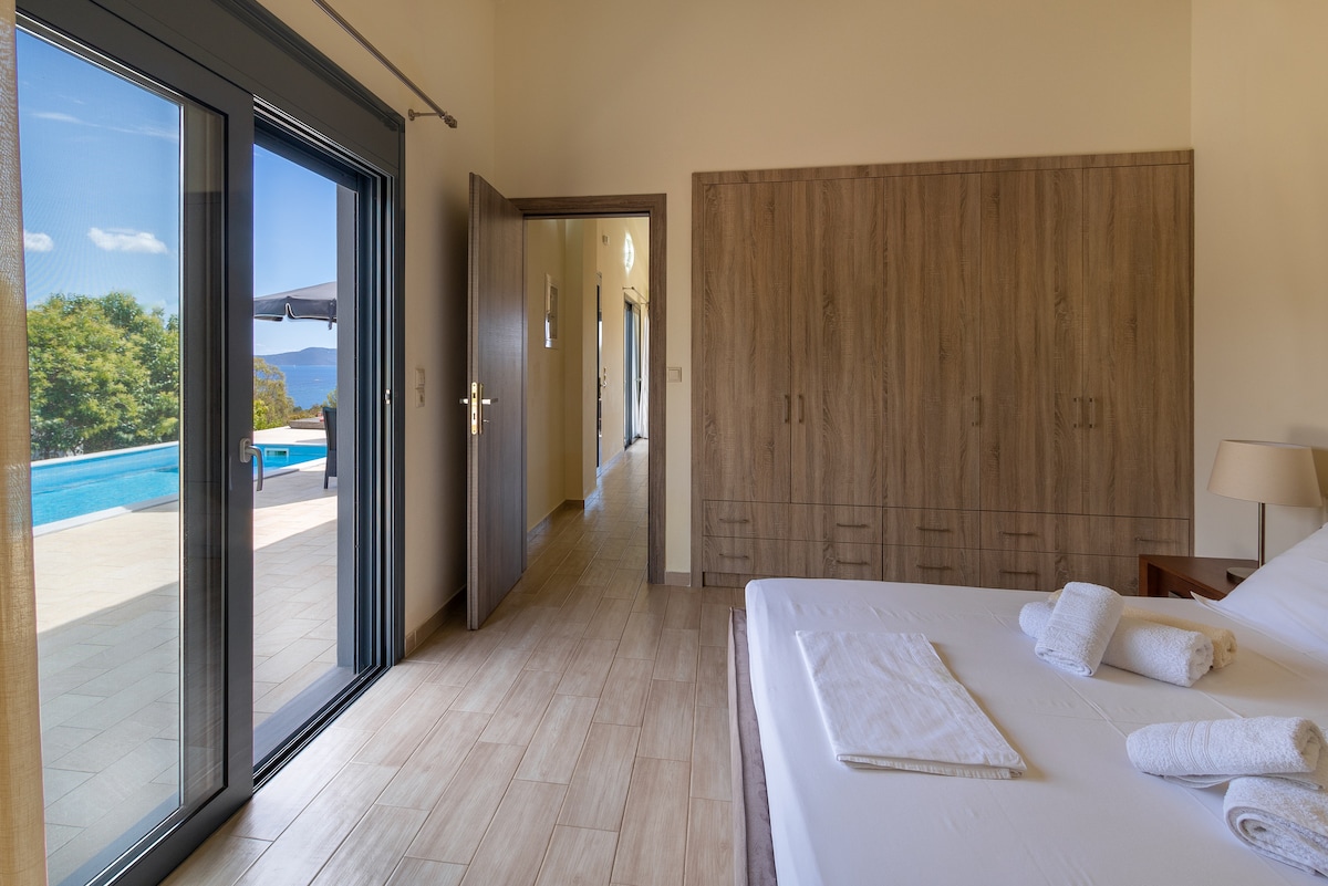 A spacious bedroom is presented with a large bed, featuring fresh towels neatly arranged. Double glass doors offer an unobstructed view of the outdoor pool area. The wooden cabinetry provides ample storage space, while natural light is welcomed from both the doors and window.