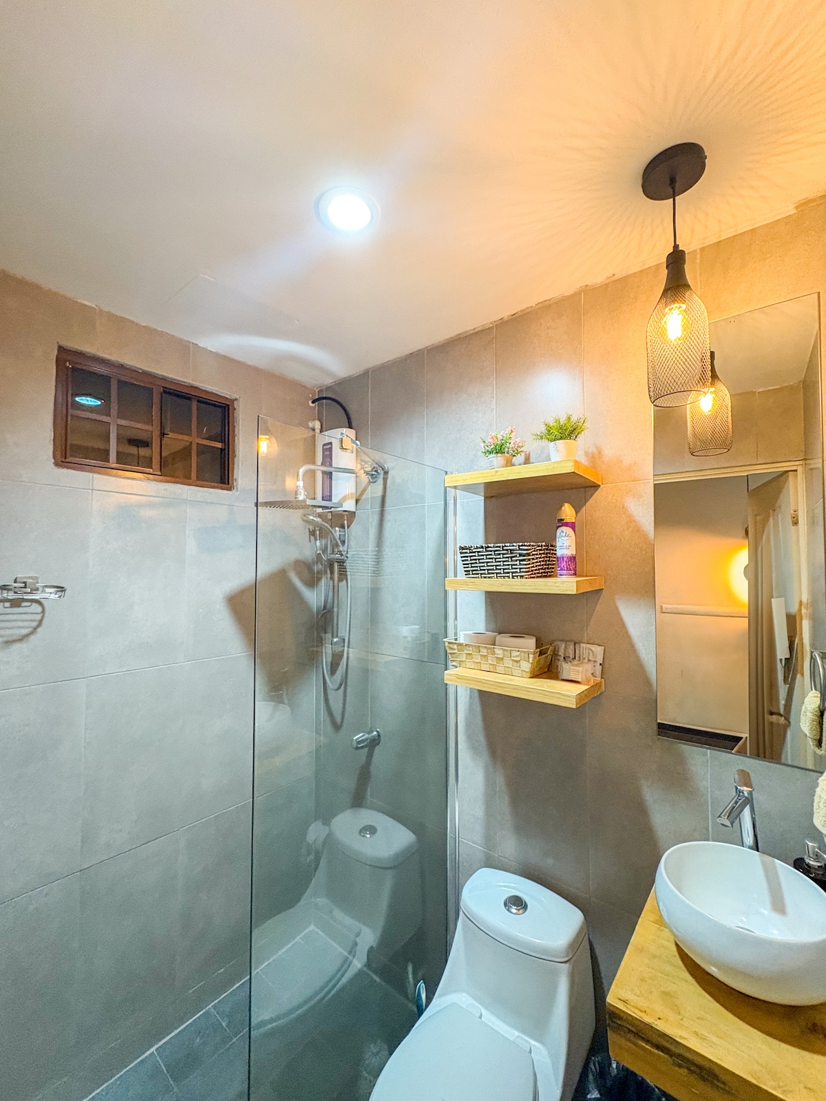 The bathroom features a glass shower enclosure with modern fixtures. A wooden shelf holds toiletries, while a circular mirror reflects light from a stylish pendant lamp. The neutral tile design is complemented by a small window, enhancing the overall spacious and clean appearance.