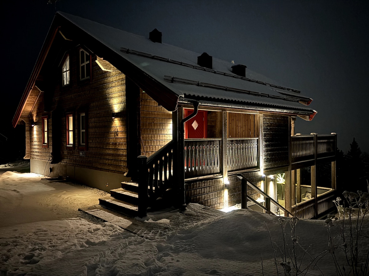 A newly built wooden mountain cabin is illuminated at night, showcasing its architectural features. The exterior is clad in timber, with a welcoming staircase leading to a furnished terrace. Snow blankets the ground, adding a serene quality to the home’s setting.