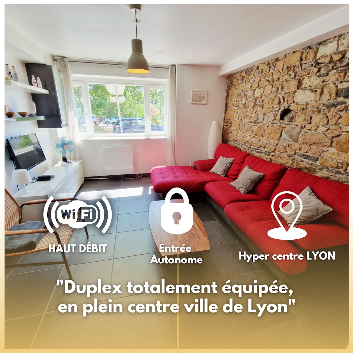 The living area features a red sectional sofa and a modern entertainment unit with a flat-screen TV. Natural light enters through a large window, illuminating the stone wall. A cozy atmosphere is created by the combination of neutral tones and the vibrant furniture.