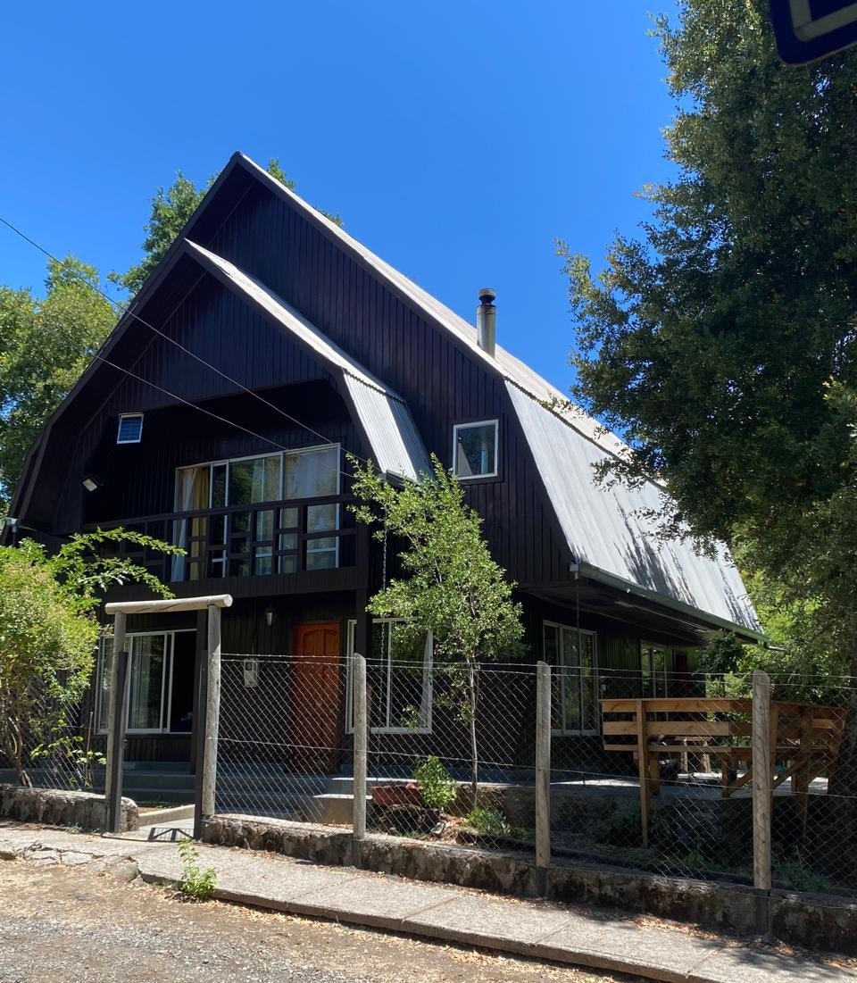 A two-story house with a distinctive sloped roof is surrounded by greenery. Large windows allow natural light to enter, while a welcoming entrance is framed by a wooden porch. A fenced yard enhances the property’s privacy in the tranquil setting.