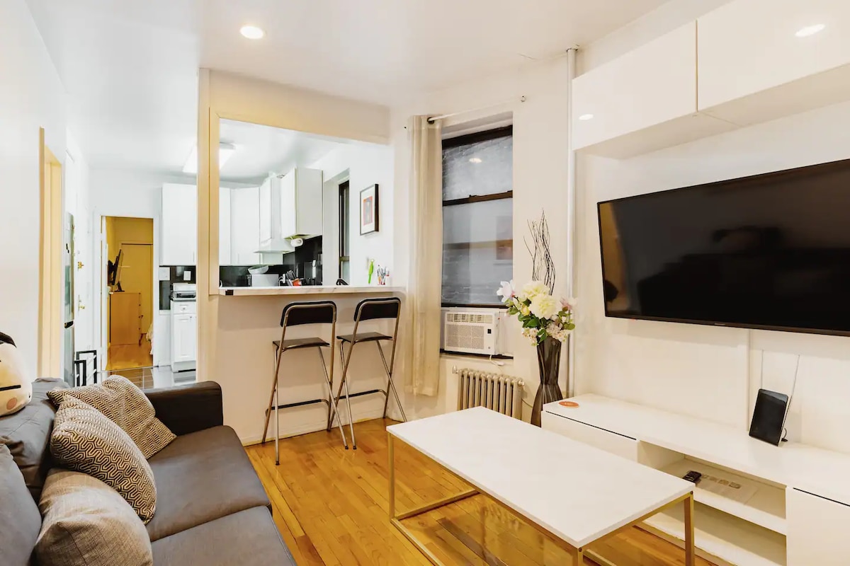 A living room area is presented with a gray sofa facing a mounted large TV. A coffee table occupies the center space, and a small dining area is visible in the background, featuring two bar stools at the kitchen counter. Natural light filters through the window.